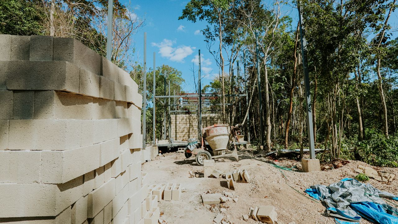 Scène de construction en extérieur avec des parpaings et un malaxeur au milieu d'un décor forestier.