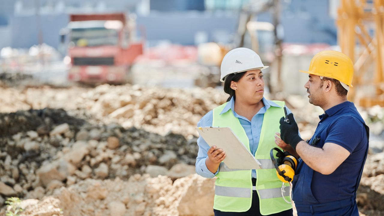 Un chef de chantier en gilet de sécurité discute avec un compagnon au pied d'un échafaudage, sur un chantier de maçonnerie en briques.