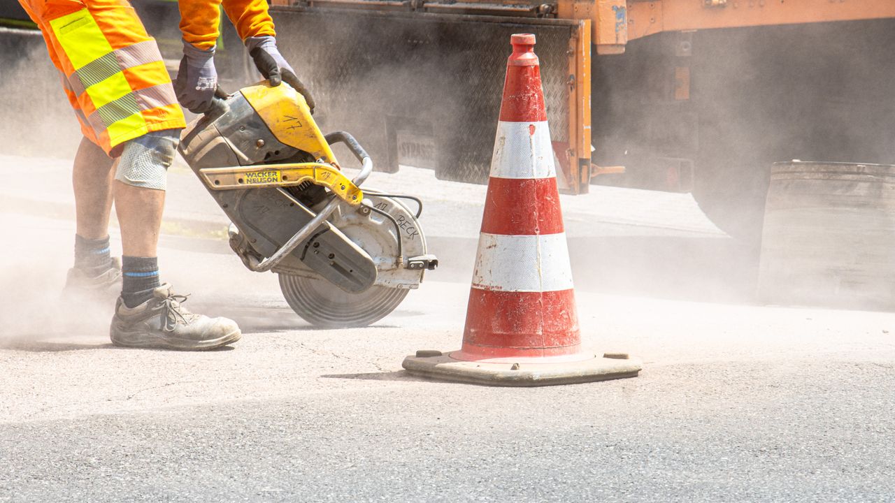 Un ouvrier du bâtiment coupe de l'asphalte avec une grande scie à côté d'un cône de signalisation dans une rue.