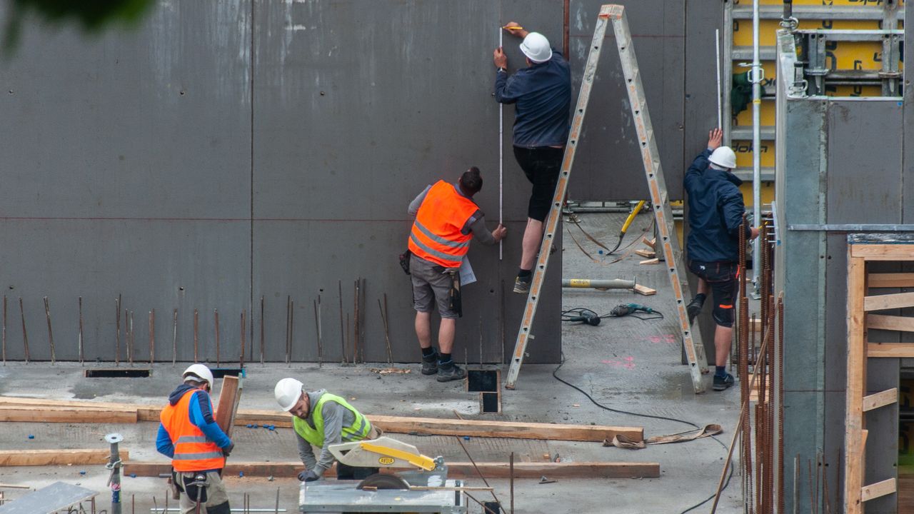 Un jeune ouvrier en gilet haute visibilité coffre un voile en béton à la tombée de la nuit, éclairé par des projecteurs de chantier, sous le regard d'un chef de chantier casqué.