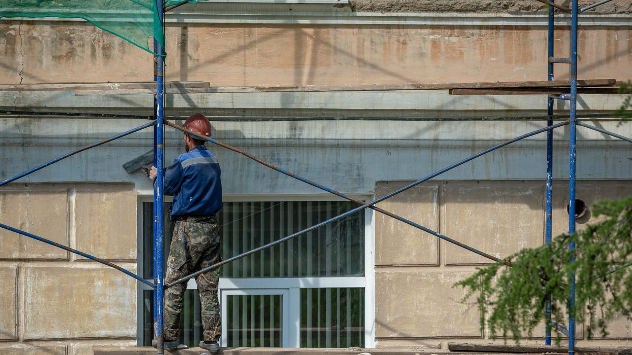 Un ouvrier du bâtiment en casque répare une façade de bâtiment sur un échafaudage dans un cadre urbain.