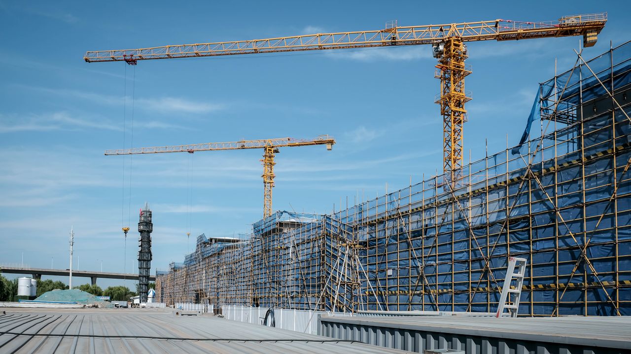 Vue d'un chantier de construction avec des grues à tour jaunes et des échafaudages sous un ciel bleu dégagé.