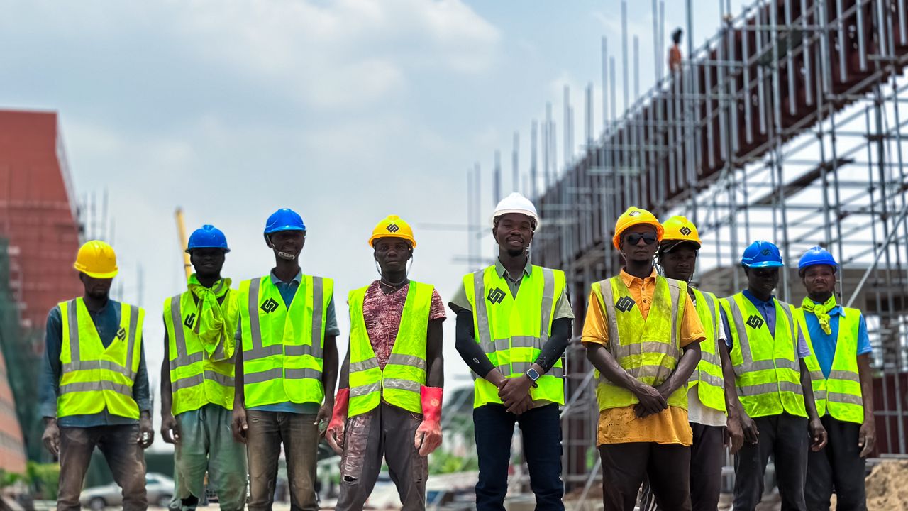 Groupe d'ouvriers en tenue de sécurité sur un chantier. Échafaudages visibles et ciel dégagé.